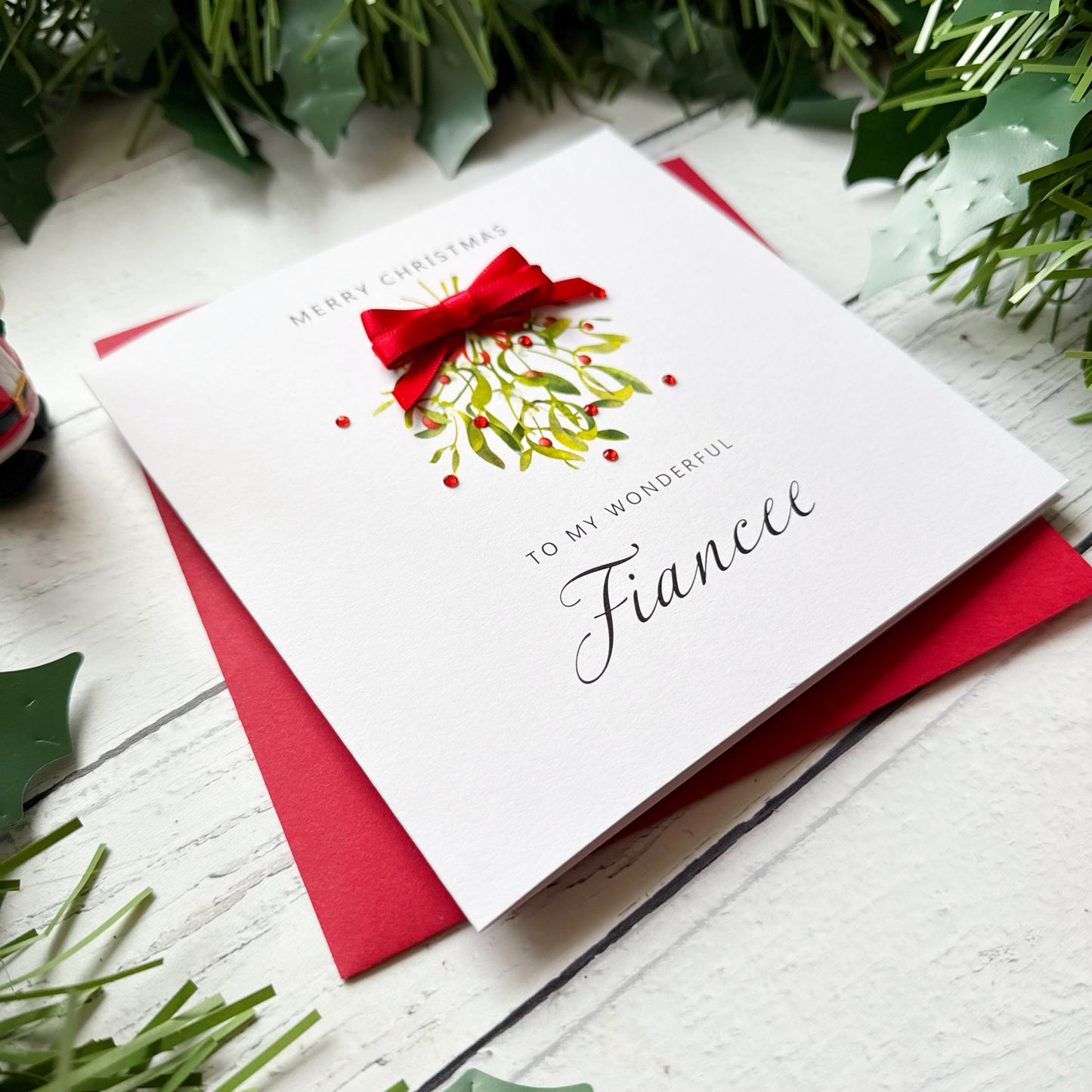 A white Christmas greeting card addressed 'To My Wonderful Fiancee' with a red bow and mistletoe illustration, placed on a red envelope over a white wooden surface with green holly leaves around it.