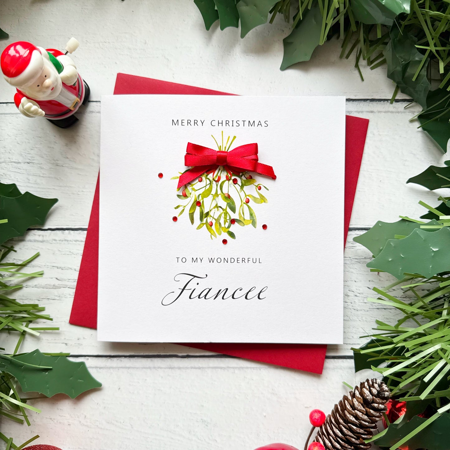 A square white Christmas card featuring a watercolour illustration of a mistletoe bunch with green leaves and red berries, tied with a real red satin ribbon bow and accented with small red gem embellishments scattered around the design. The text "Merry Christmas" is printed in small spaced capitals at the top of the card, with "to my Wonderful" in small capitals and "Fiancee" in large elegant black cursive script below the illustration. The card is displayed at an angle on a matching red envelope on a white