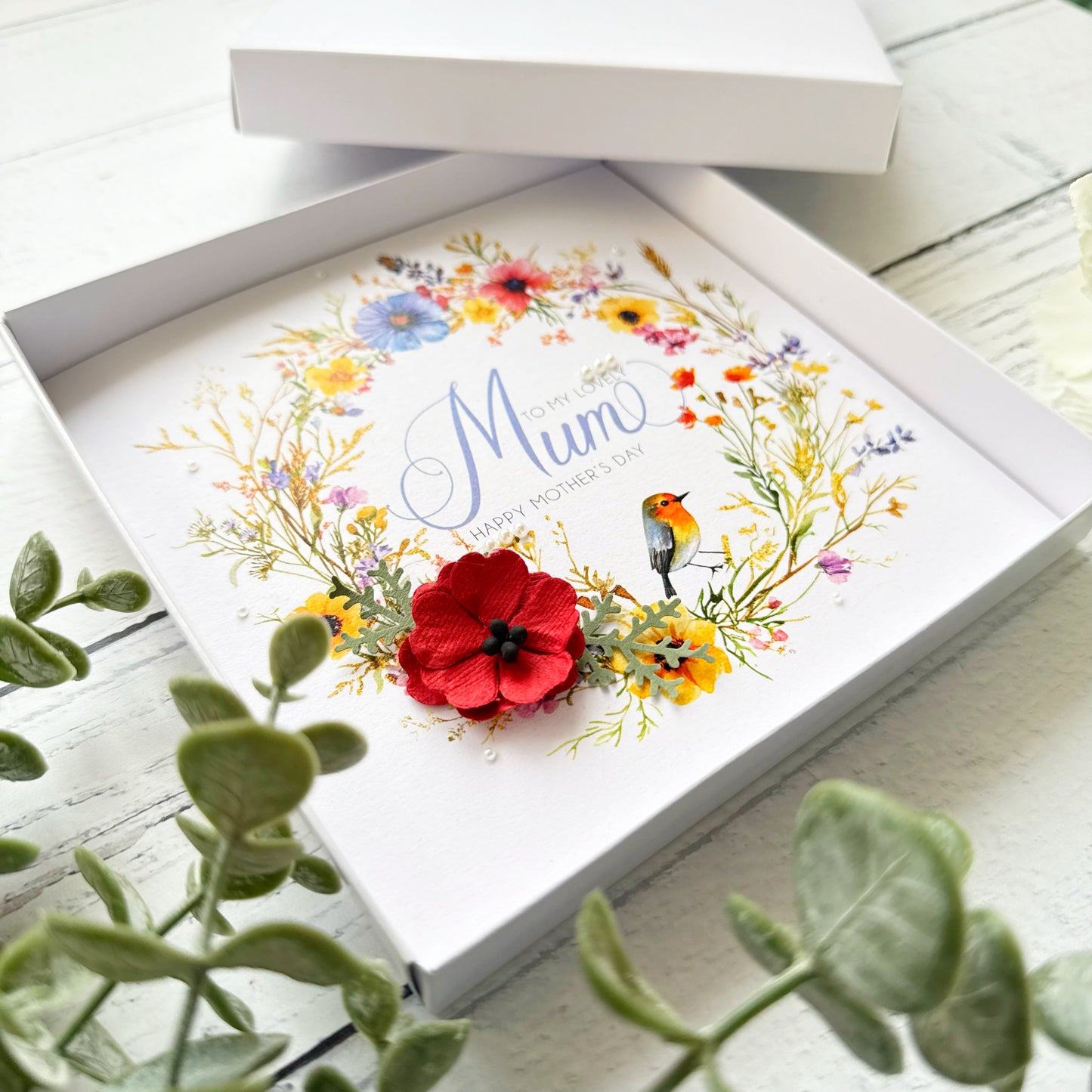 Greeting card with floral design and 'Mum' text, placed inside an open box on a wooden surface.