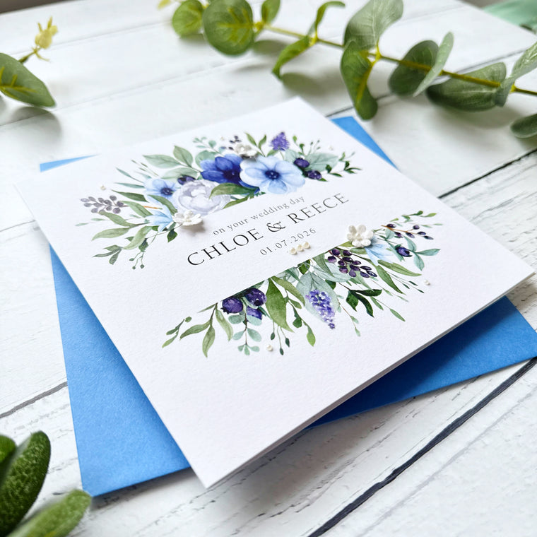 Wedding card with blue floral design decorated with pearls and paper flowers. Featuring personalised text details and a blue envelope on a white wooden surface