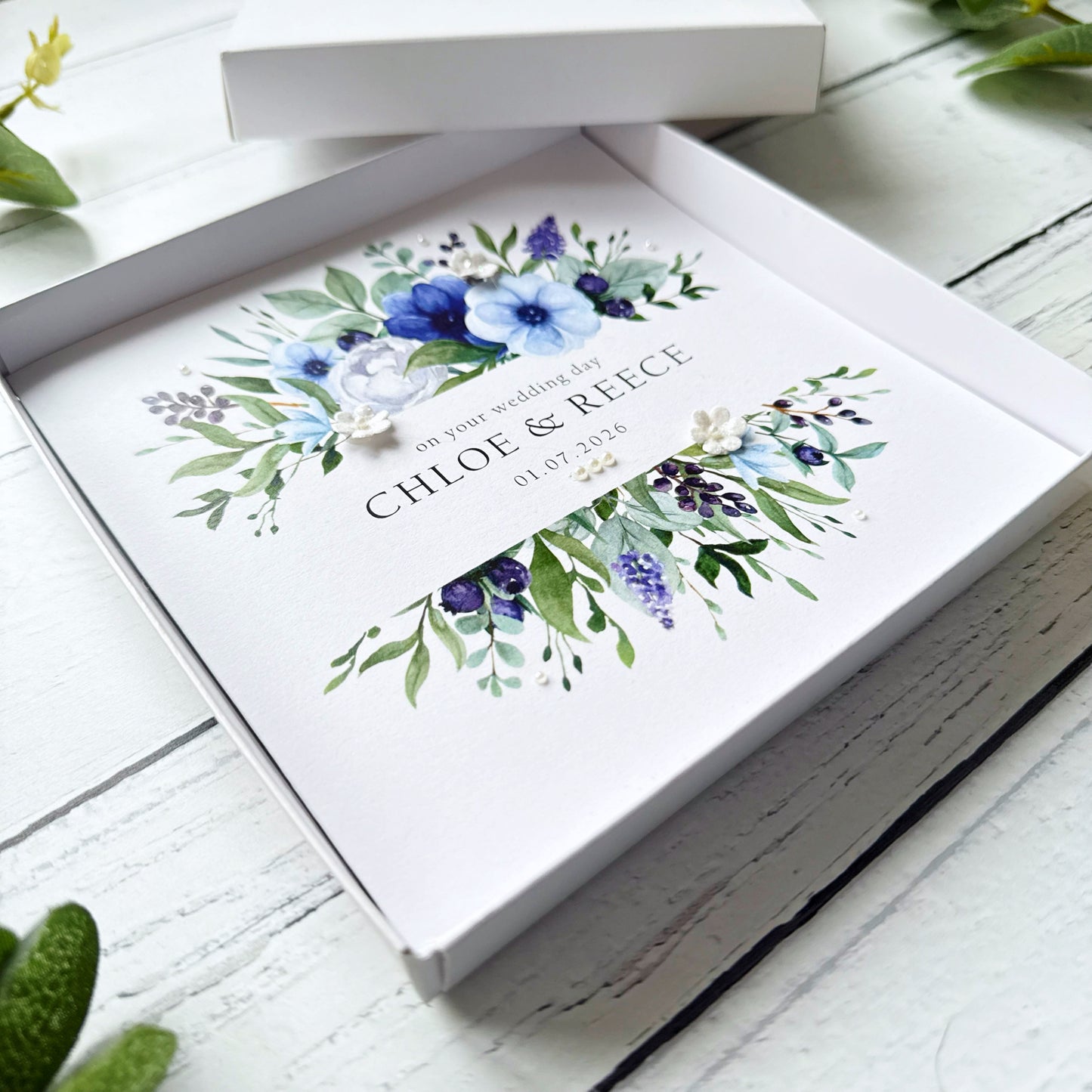 Wedding card with blue floral design and personalised text decorated with tiny pearls and paper flowers in a white box.