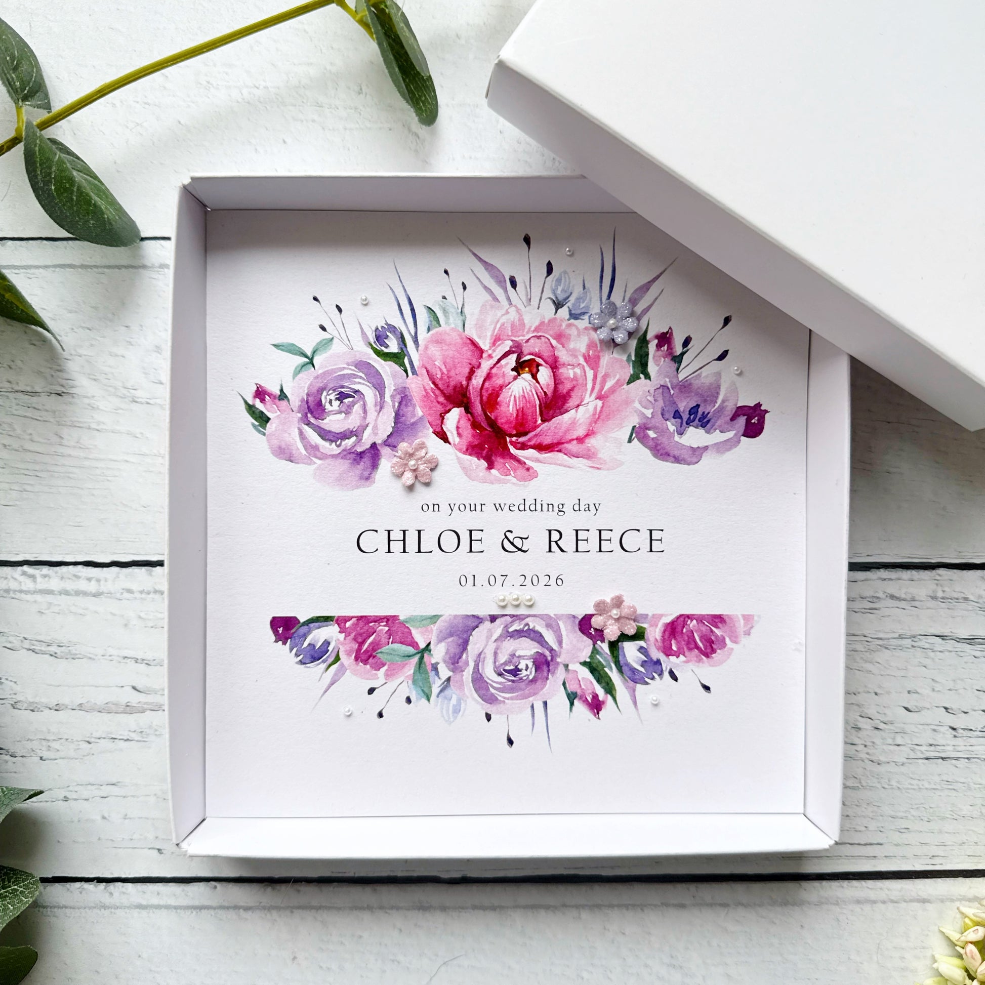 Decorative wedding card with pink and purple floral design and personalised text. Decorated with tiny pearls and paper flowers, in a box on a white wooden surface