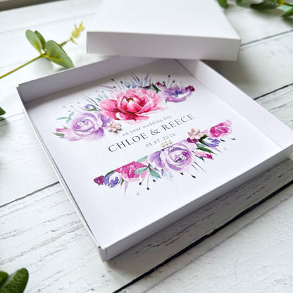 Pink and Purple Floral design wedding card decorated with tiny pearls and 3 paper flowers in a box on a white wooden surface