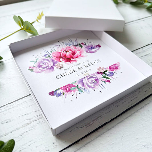 Pink and Purple Floral design wedding card decorated with tiny pearls and 3 paper flowers in a box on a white wooden surface