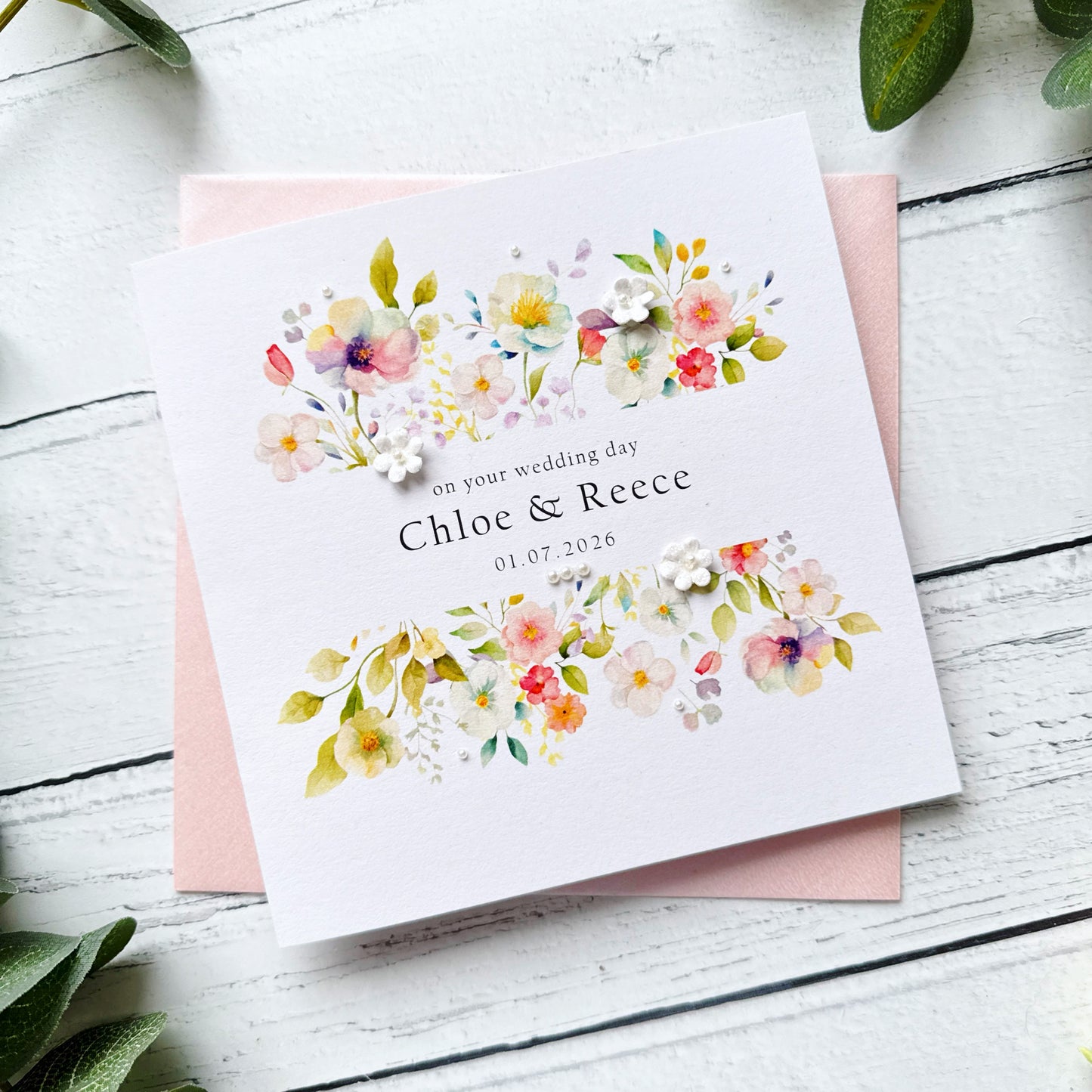 Floral wedding card with personalised text, pearls and paper flowers on a pink envelope on a white wooden surface