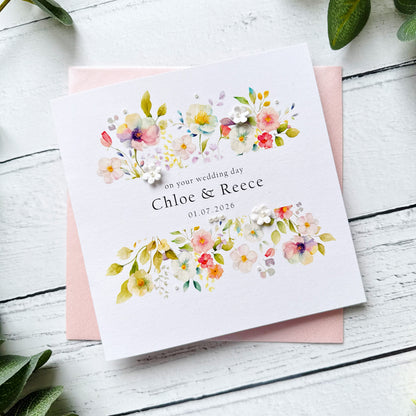Floral wedding card with personalised text, pearls and paper flowers on a pink envelope on a white wooden surface