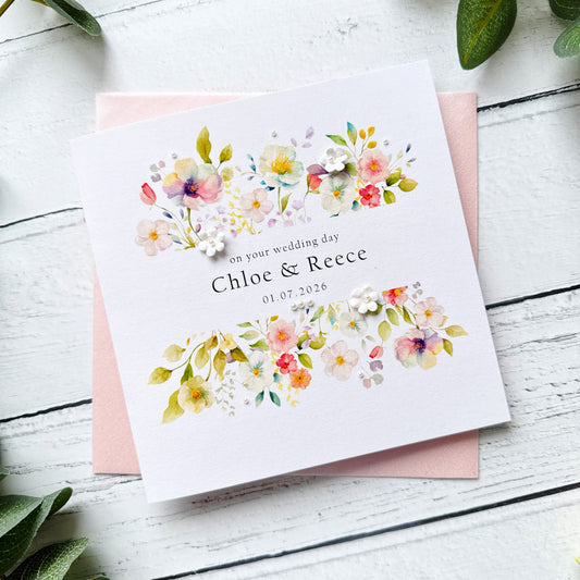 Floral wedding card with personalised text, pearls and paper flowers on a pink envelope on a white wooden surface