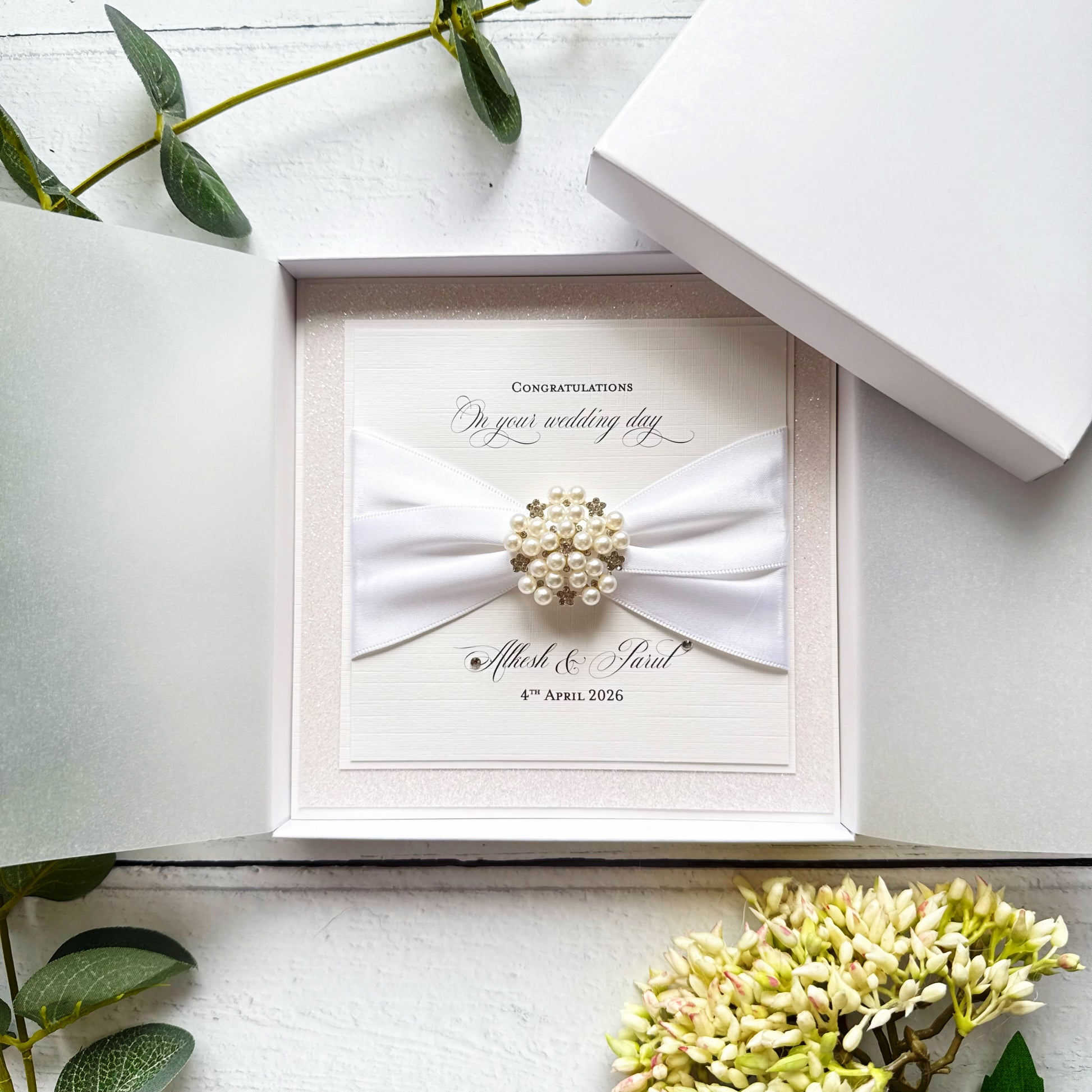 Wedding card with a bow and brooch in an open box on a white surface with greenery.