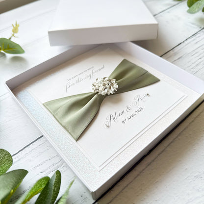 Wedding card with a green bow and brooch in an open box on a wooden surface.