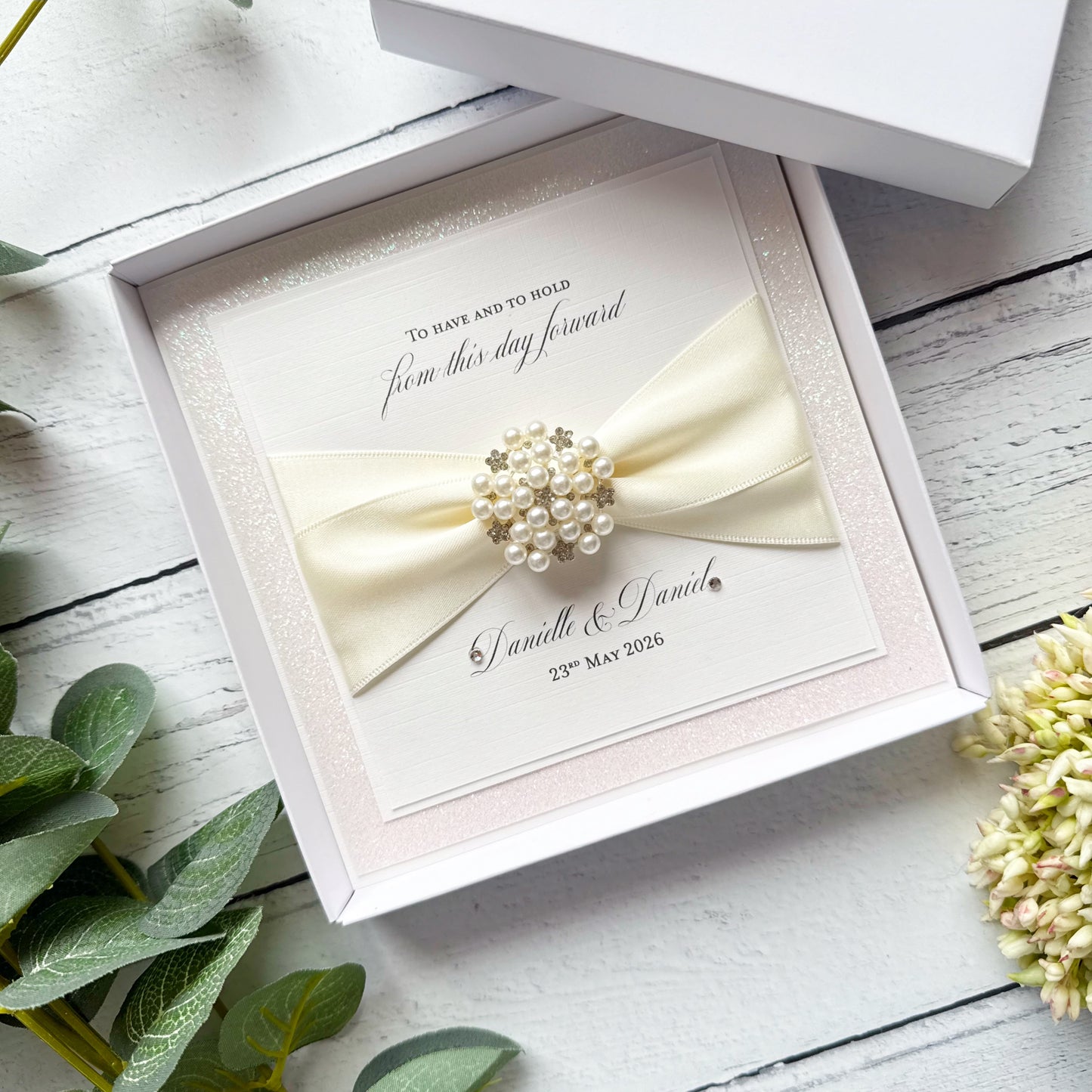 PersonaliSed wedding card with a bow and brooch in a decorative box on a wooden surface.