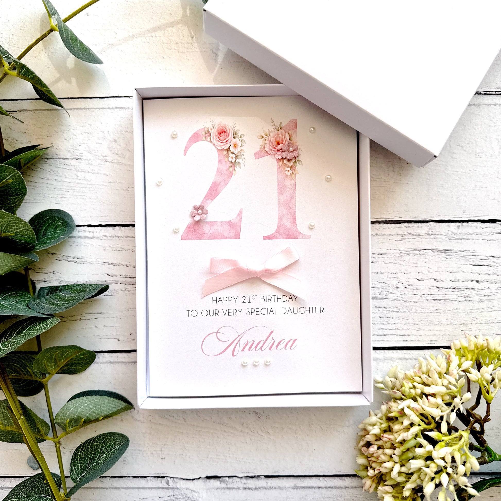 21st birthday card with floral design with a bow and pearls. With personalised text on a wooden surface with plants.