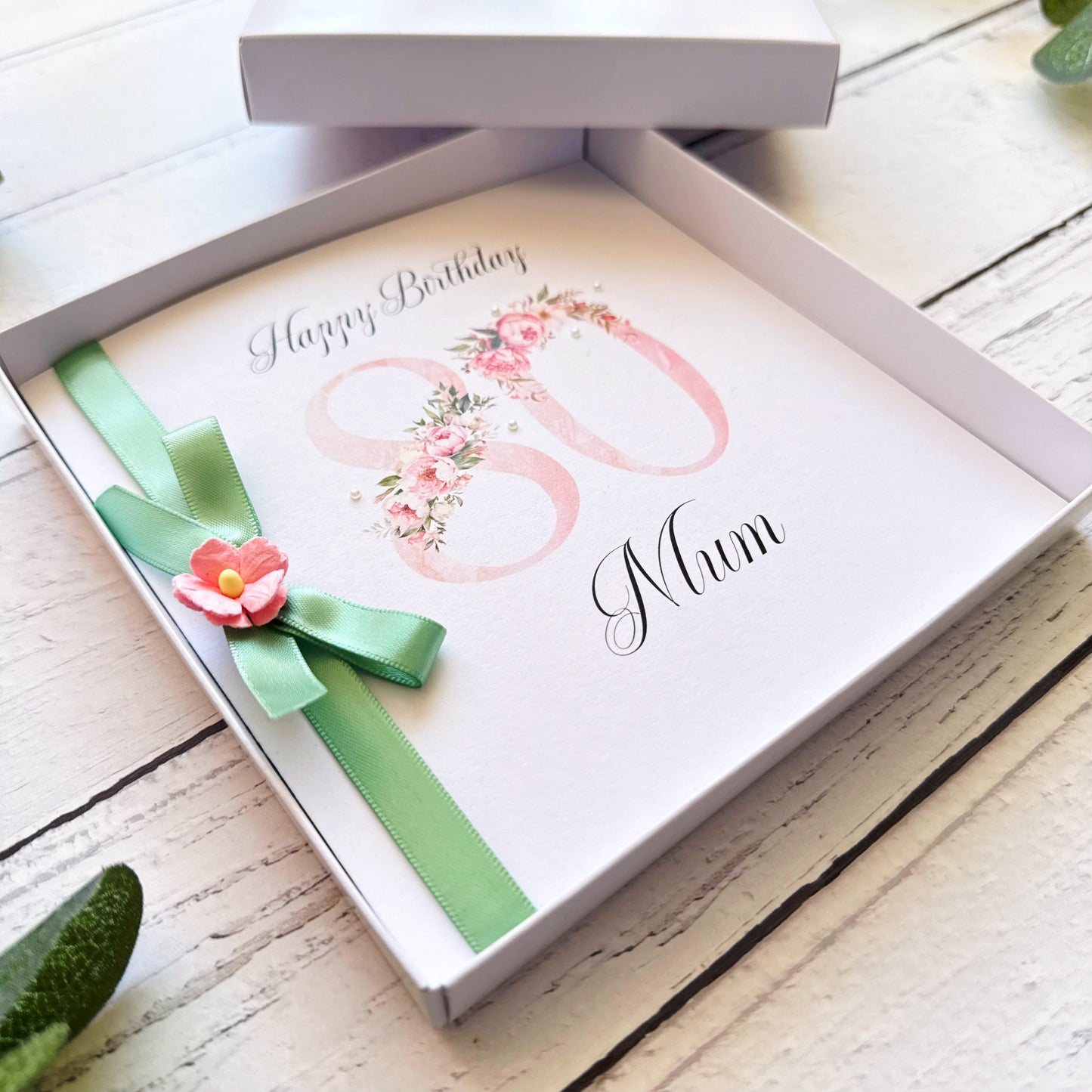Greeting card with floral design and 'Happy Birthday (name)' text in a box on a wooden surface.
