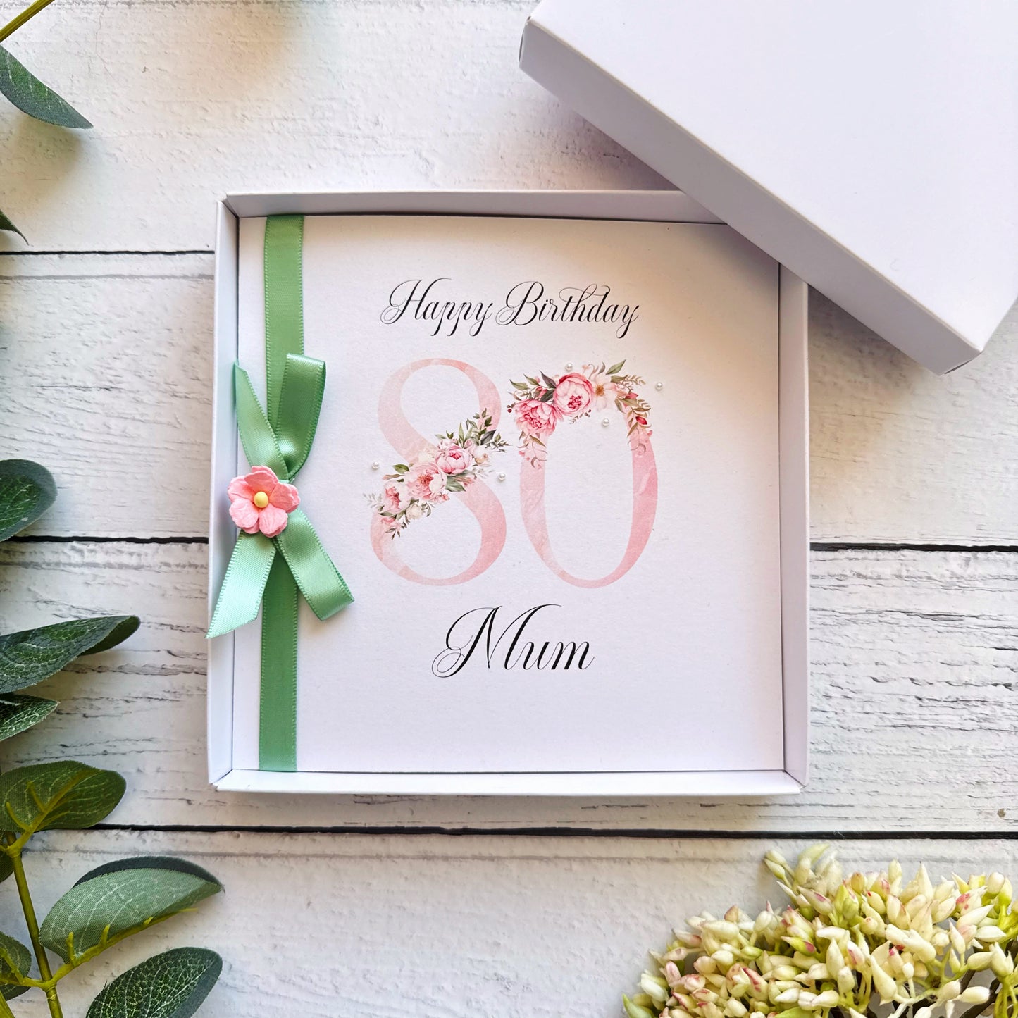 Greeting card with 'Happy Birthday 80 Mum' text, green ribbon, and flowers on a wooden surface.