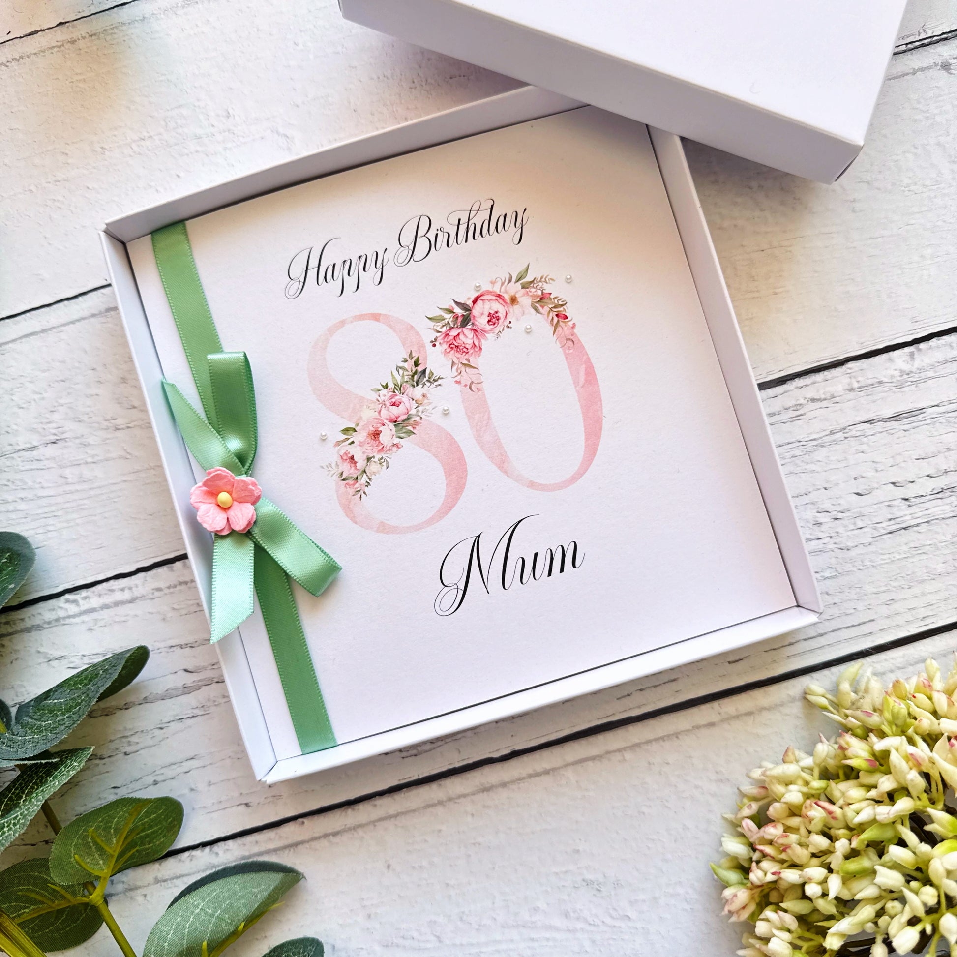 80th birthday card for 'Mum' with floral design and green ribbon in a box on a wooden surface.