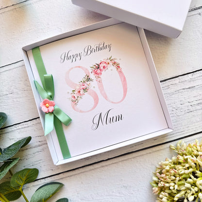 80th birthday card for 'Mum' with floral design and green ribbon in a box on a wooden surface.