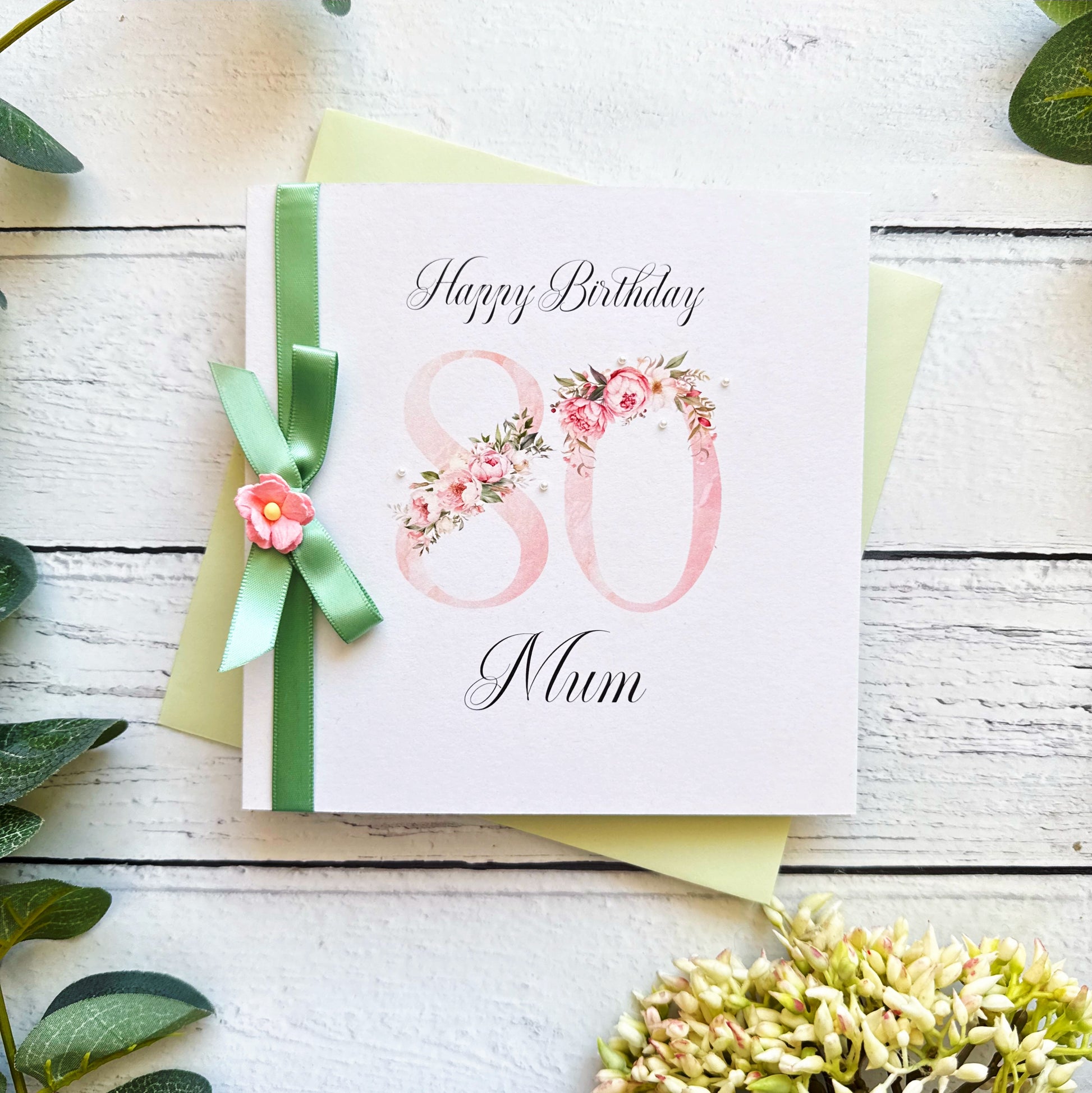 80th birthday card for mum with floral design and green ribbon on a wooden surface.