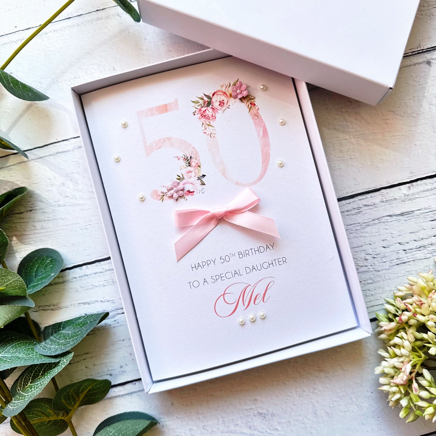 Personalised greeting card with floral numbers design hand-finished with a bow, paper flowers and pearls on a wooden surface with greenery.