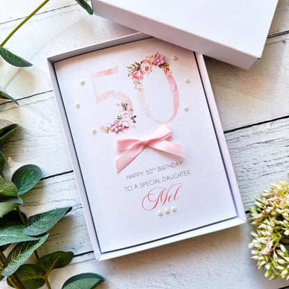 Personalised greeting card with floral numbers design hand-finished with a bow, paper flowers and pearls on a wooden surface with greenery.