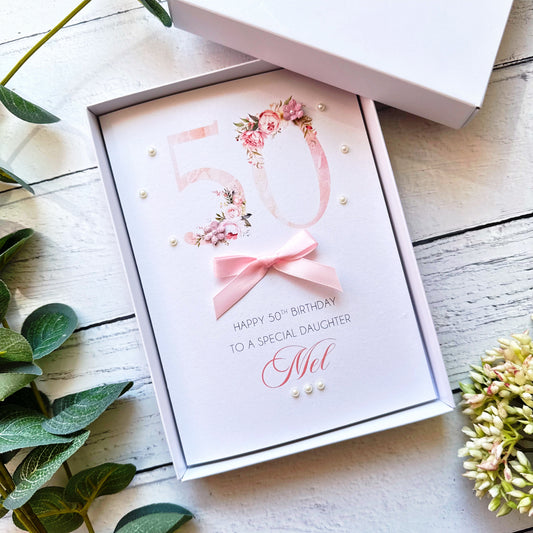 Personalised greeting card with floral numbers design hand-finished with a bow, paper flowers and pearls on a wooden surface with greenery.