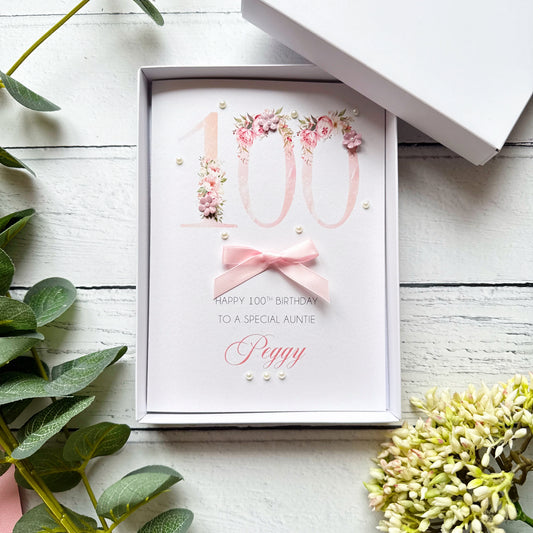 100th birthday greeting card with floral number design and personalised birthday message. Hand-finished with a pink bow, pearls and paper flowers on a white wooden surface with greenery.