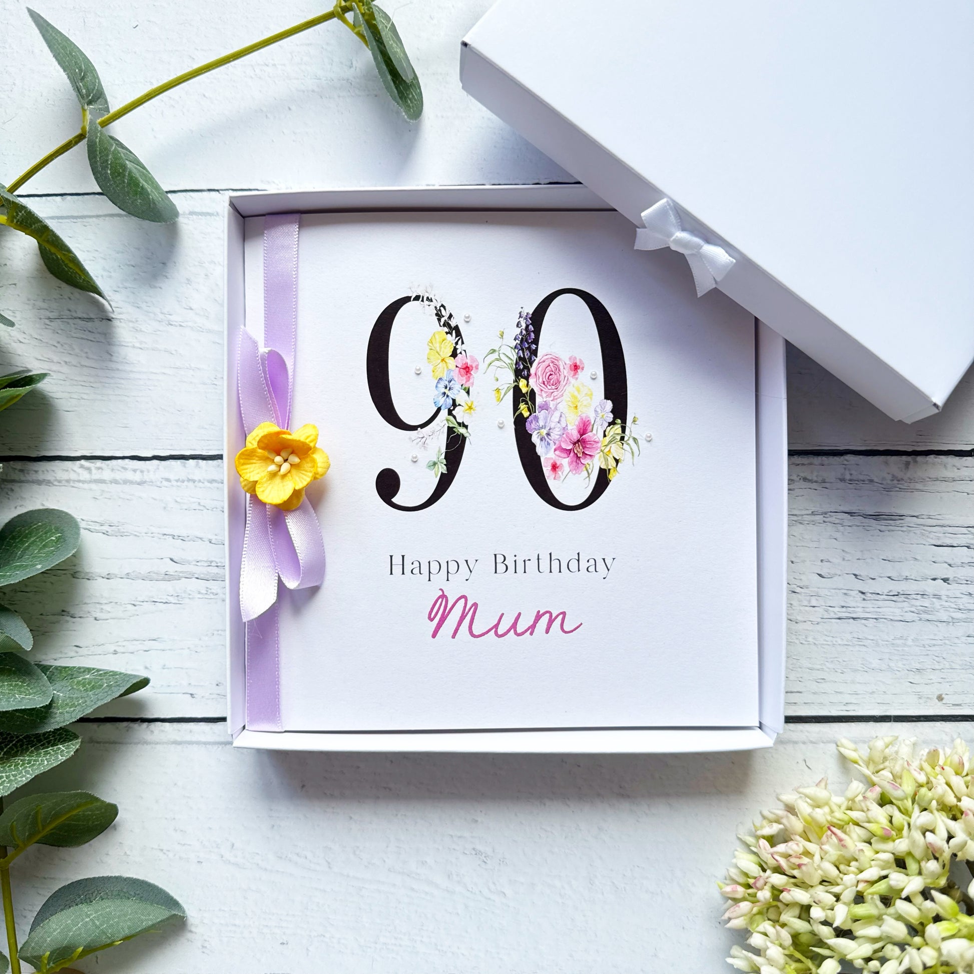 90th birthday Greeting card with 'Happy Birthday Mum'. Decorated with lilac ribbon yellow flower and pearls on a white wooden surface with decorative elements.