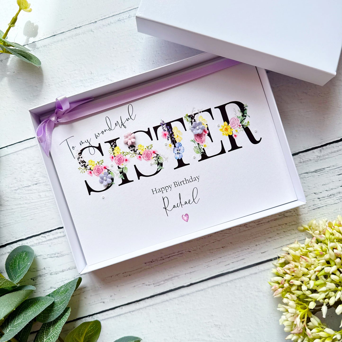 Greeting card with floral lettering for a sister, in a box, in shades of purple, pink and yellow on a white wooden surface