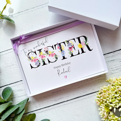 Greeting card with floral lettering for a sister, in a box, in shades of purple, pink and yellow on a white wooden surface