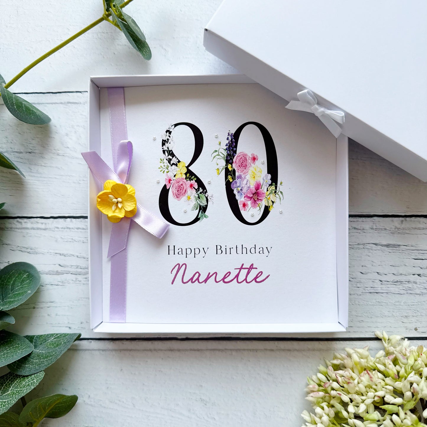 80th Birthday Greeting card with floral 80 Happy Birthday (name). With lilac bow, yellow paper flower and tiny pearls on a white background with decorative elements.