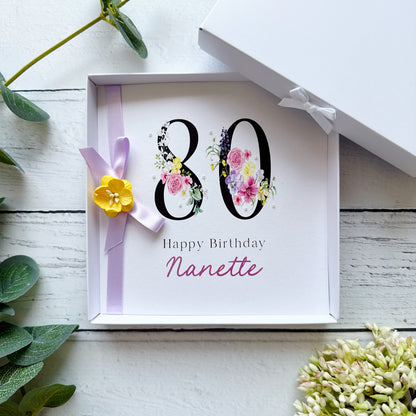 80th Birthday Greeting card with floral 80 Happy Birthday (name). With lilac bow, yellow paper flower and tiny pearls on a white background with decorative elements.
