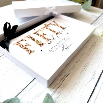 Greeting card with leopard print text "Fifty and Fabulous" 'Happy Birthday' in a white box on a wooden surface.