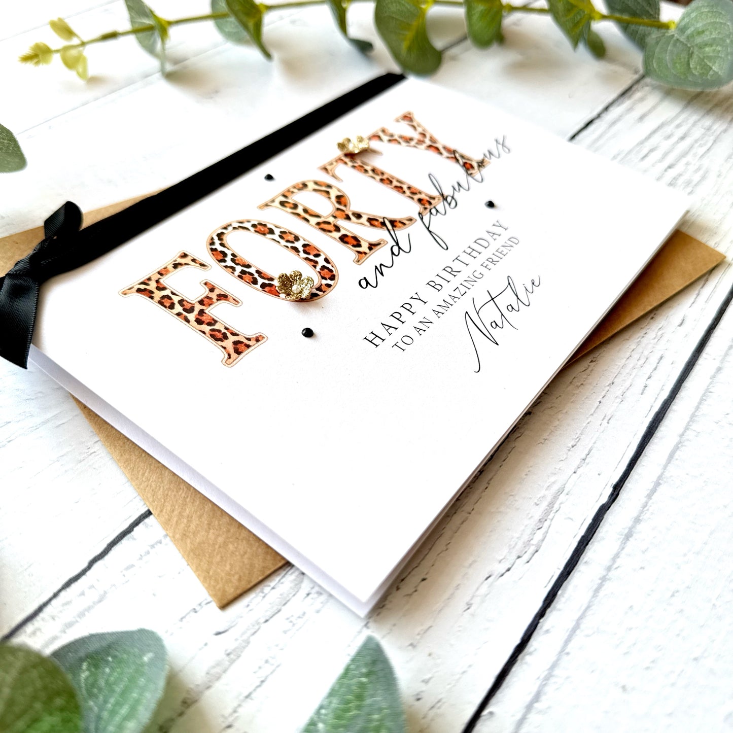 Birthday card with leopard print text "forty and fabulous" and black bow on a white wooden surface with greenery.