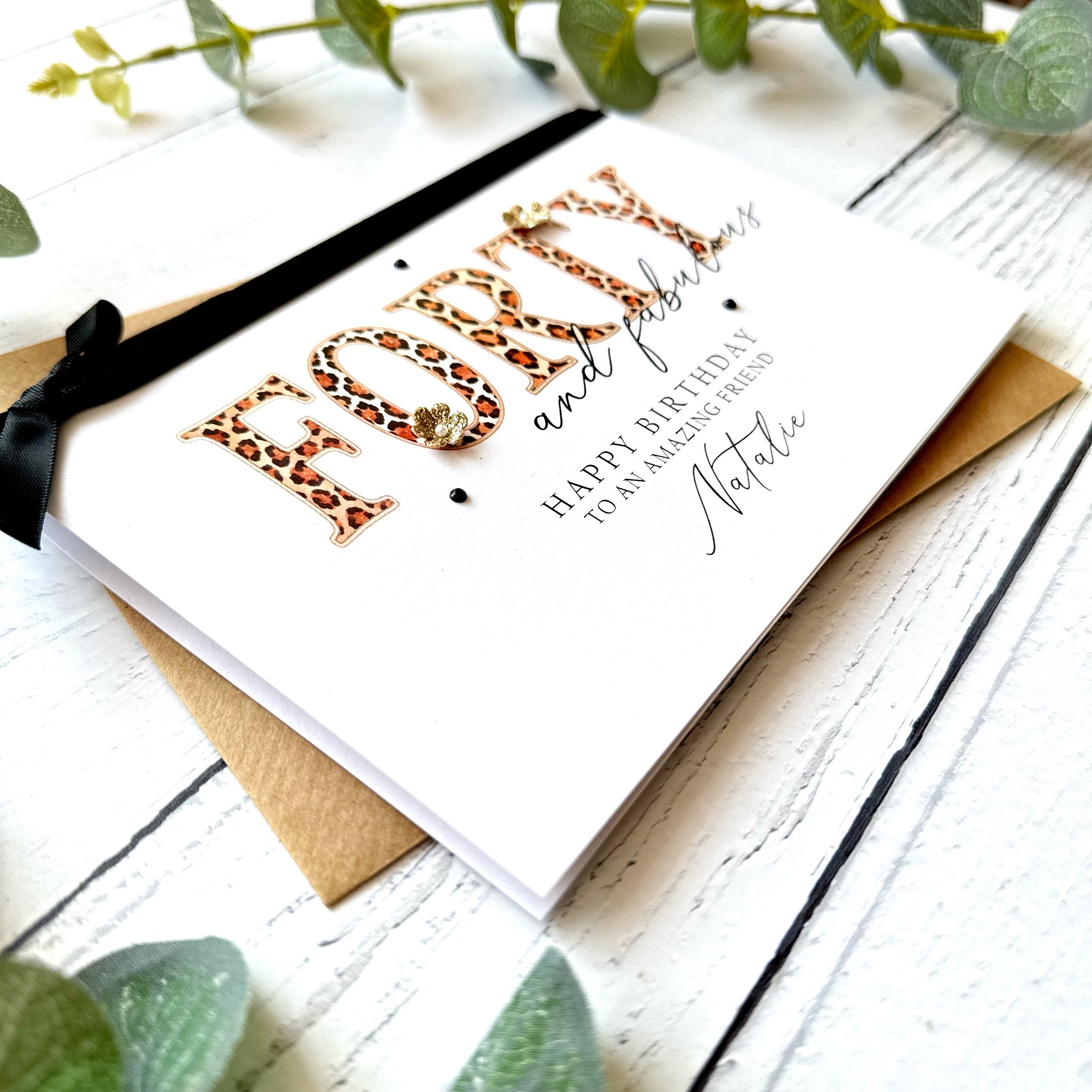 Birthday card with leopard print text "forty and fabulous" and black bow on a white wooden surface with greenery.