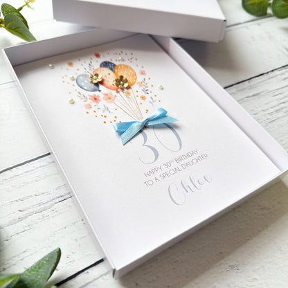 Greeting card with balloons and a bow, paper flowers and pearls with personalised text "happy 30th birthday to a special daughter (name) in an open box on a wooden surface