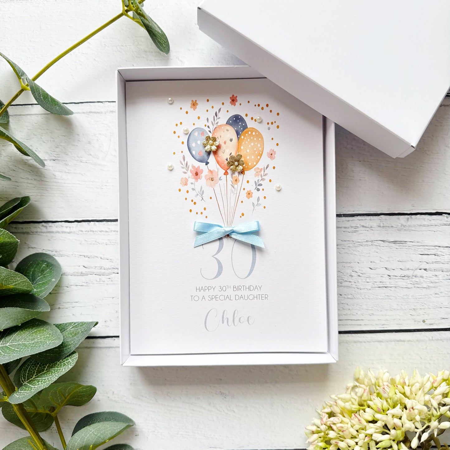 30th birthday greeting card with balloons and a bow. With personalised text in an open box on a wooden surface.