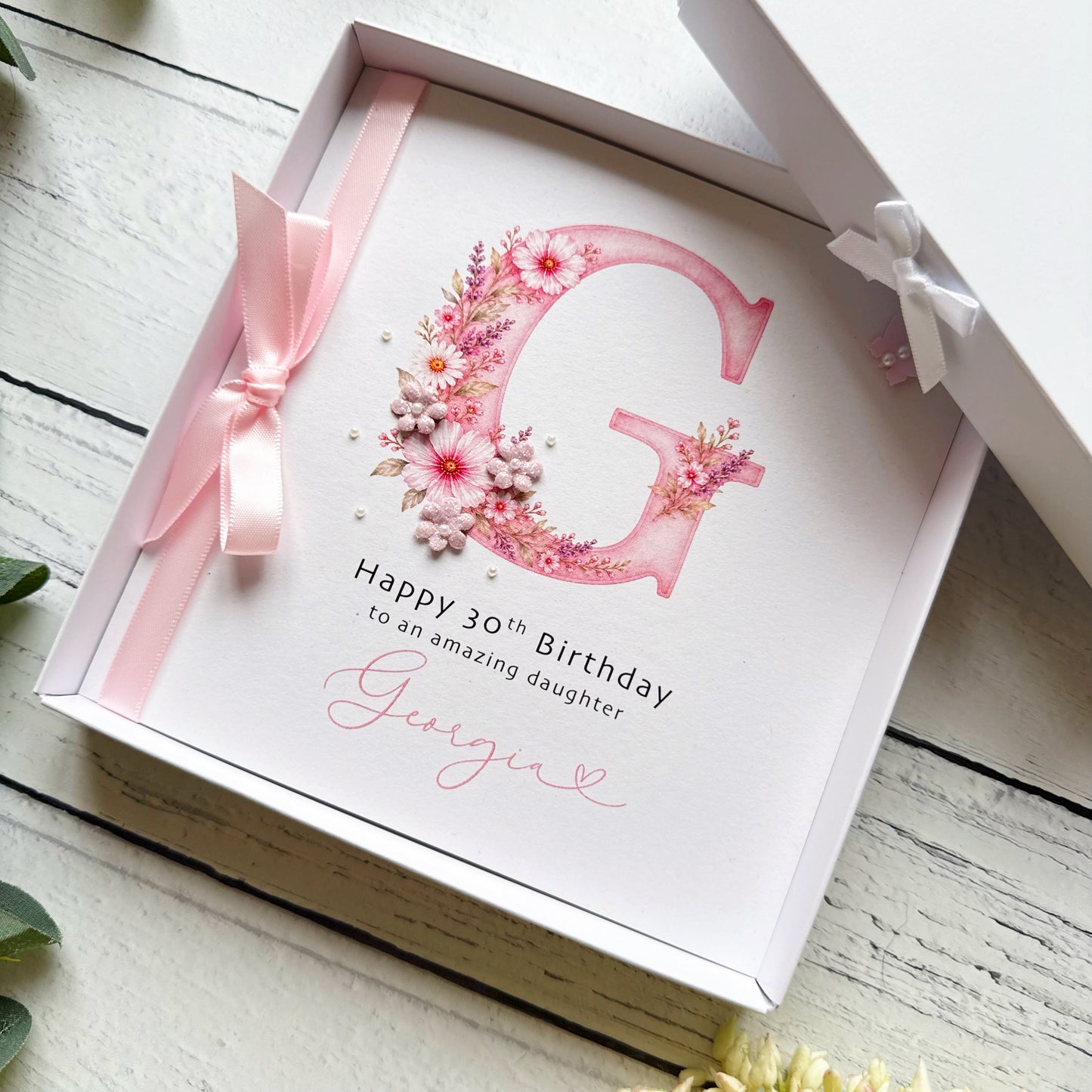 Greeting card with pink monogram floral design and personalised details in a decorative box. Decorated with a ribbon, paper flowers and tiny pearls on a wooden surface.