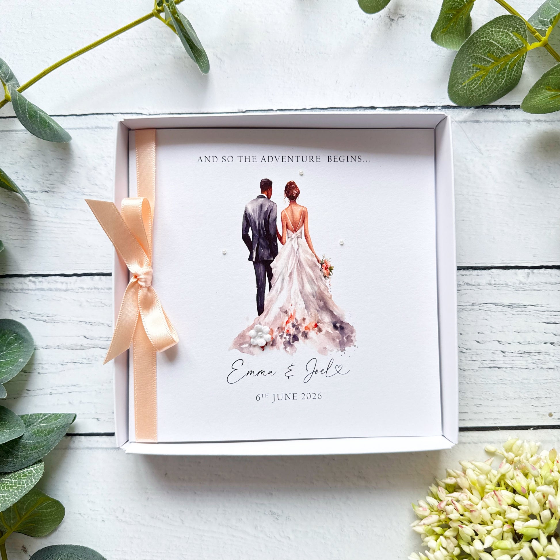 White wedding card with interracial couple, black groom white bride. Text reads "and so the adventure begins" followed by the couples names and wedding date. Decorated with a ribbon, small paper flower and tiny pearls