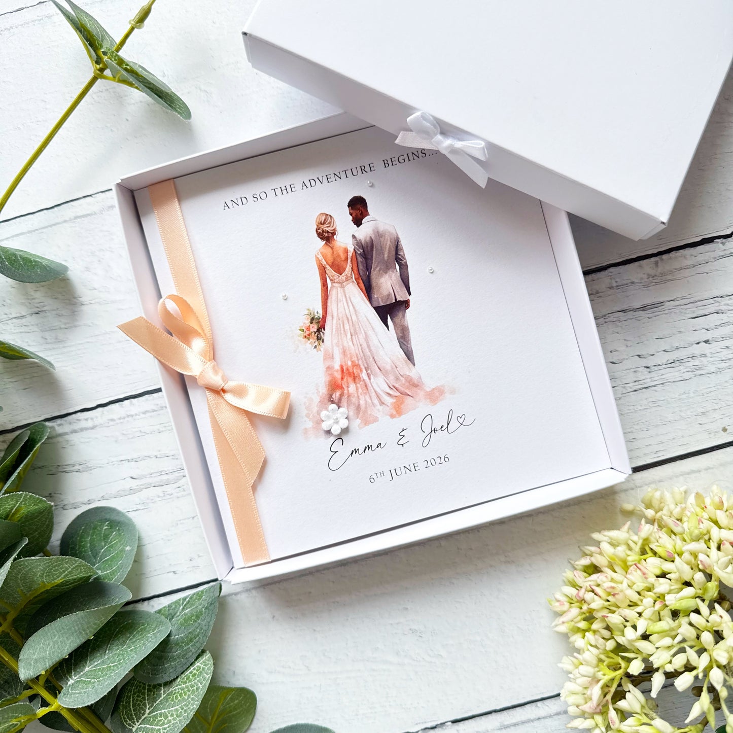 White wedding card with interracial couple, black groom white bride. Text reads "and so the adventure begins" followed by the couples names and wedding date. Decorated with a ribbon, small paper flower and tiny pearls