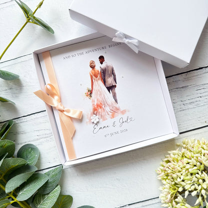 White wedding card with interracial couple, black groom white bride. Text reads "and so the adventure begins" followed by the couples names and wedding date. Decorated with a ribbon, small paper flower and tiny pearls