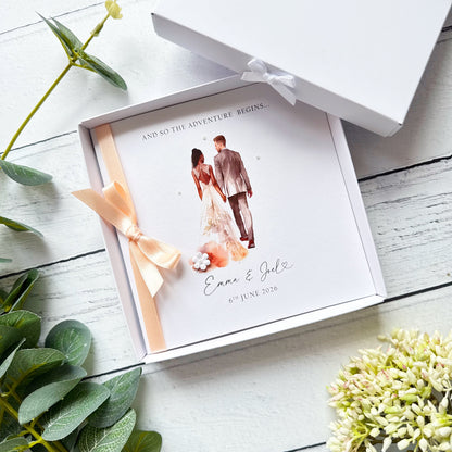 White wedding card with interracial couple, white groom black bride. Text reads "and so the adventure begins" followed by the couples names and wedding date. Decorated with a ribbon, small paper flower and tiny pearls