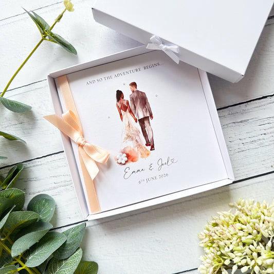 White wedding card with interracial couple, white groom black bride. Text reads "and so the adventure begins" followed by the couples names and wedding date. Decorated with a ribbon, small paper flower and tiny pearls