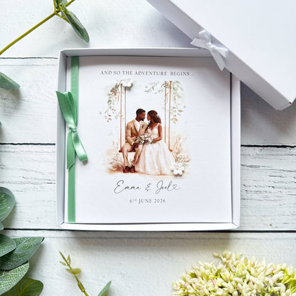 White wedding card with black bride and groom. Text reads "and so the adventure begins" followed by the couples names and wedding date. Decorated with a ribbon, small paper flower and tiny pearls