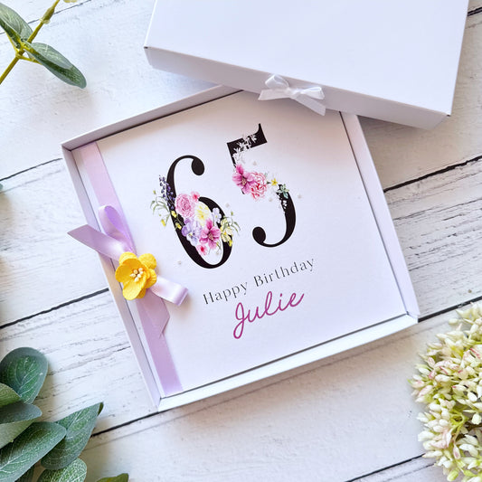 A square white greeting card displaying a large bold "65" decorated with watercolour flowers including pink roses, purple pansies, lavender and yellow blooms. Below the number reads "Happy Birthday" in black serif font, with the name "Sarah" written in pink cursive script. The card is tied with a lilac satin ribbon and finished with a small handmade yellow flower. It sits in presentation box, styled on a white painted wooden surface surrounded by green eucalyptus leaves and white floral sprigs
