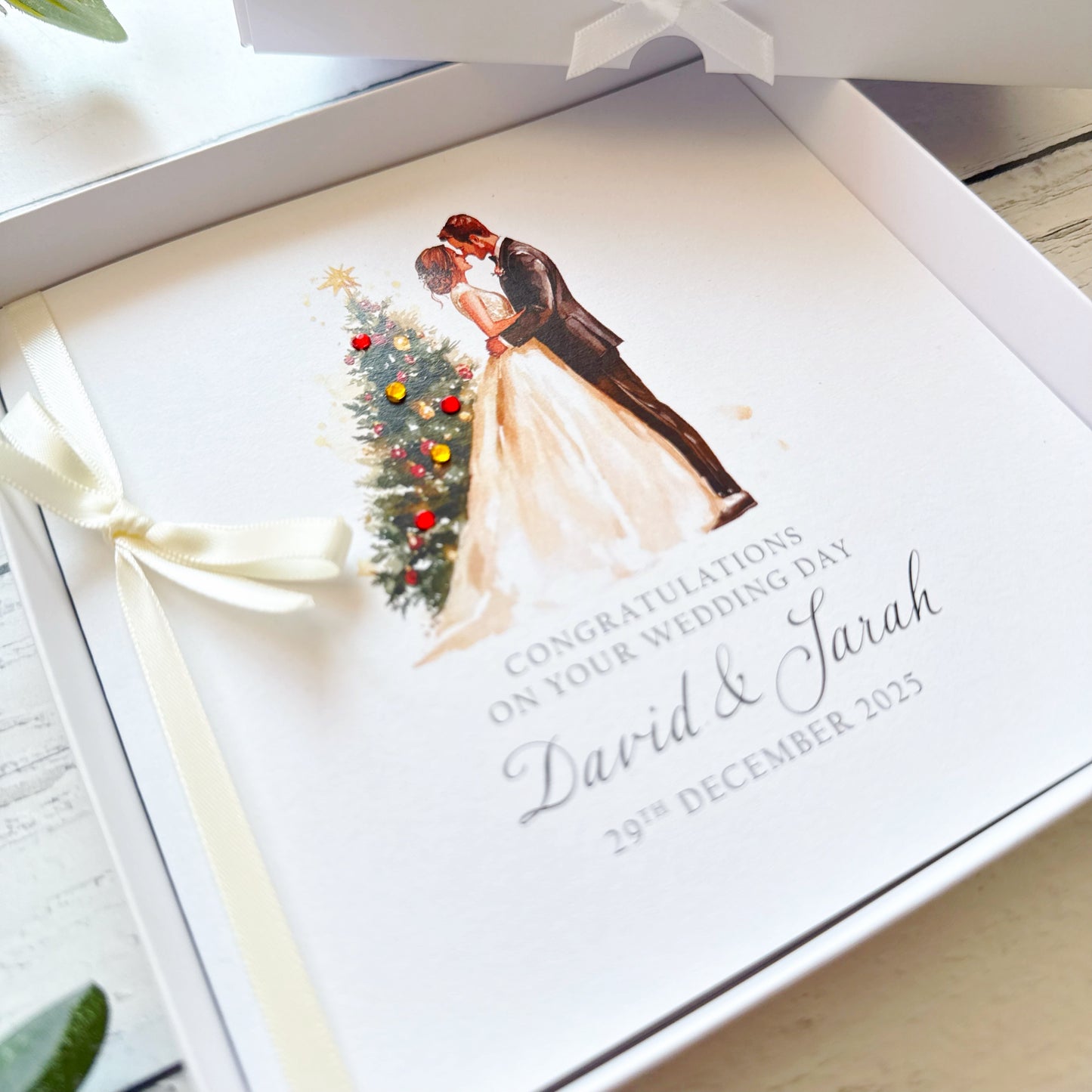 Elegant personalised wedding card featuring a bride and groom standing beside a decorated Christmas tree in a soft watercolour style. The card includes customised names and wedding date details and is finished with a cream ribbon bow. Displayed on a rustic white wooden background with greenery and floral accents.
