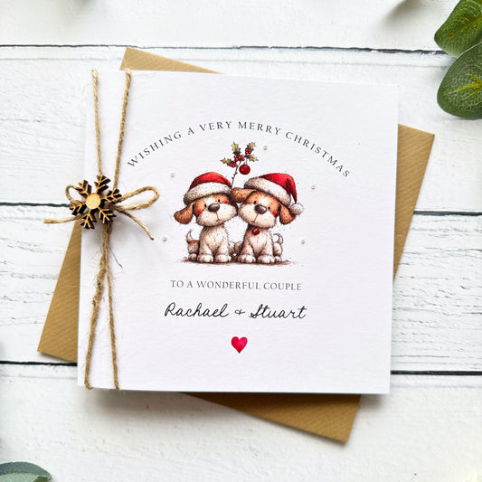 A square white Christmas card featuring two fluffy dogs in red Santa hats beneath a holly sprig, with pearl embellishments and red glitter details. Text reads "Wishing a Very Merry Christmas to a Wonderful Couple" with "Rachael & Stuart" in cursive and a red heart. Tied with natural twine and a wooden snowflake charm, displayed on a kraft envelope with candy canes and red berry sprigs.