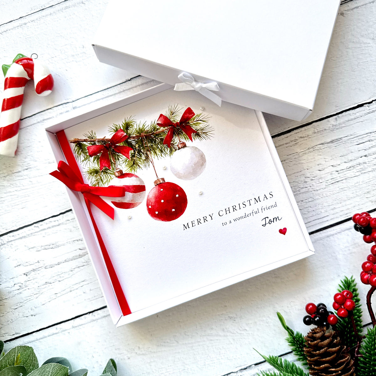 Personalised Christmas card featuring watercolour red and white baubles hanging from evergreen fir branches decorated with red bows. The branches are hand-finished with sparkling glitter to create a snowy effect. The card has a red satin ribbon bow, tiny pearl embellishments and personalised wording reading Merry Christmas to a wonderful friend Tom. Supplied with a red envelope, with an optional white presentation box.