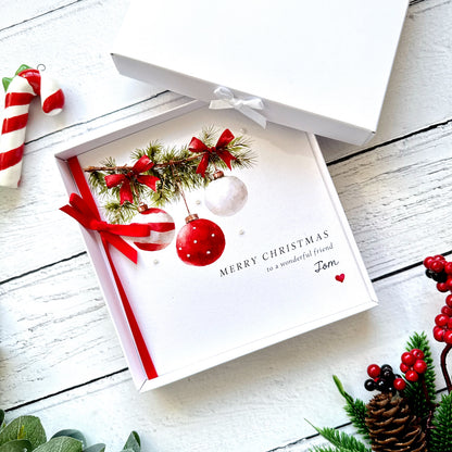 Personalised Christmas card featuring watercolour red and white baubles hanging from evergreen fir branches decorated with red bows. The branches are hand-finished with sparkling glitter to create a snowy effect. The card has a red satin ribbon bow, tiny pearl embellishments and personalised wording reading Merry Christmas to a wonderful friend Tom. Supplied with a red envelope, with an optional white presentation box.