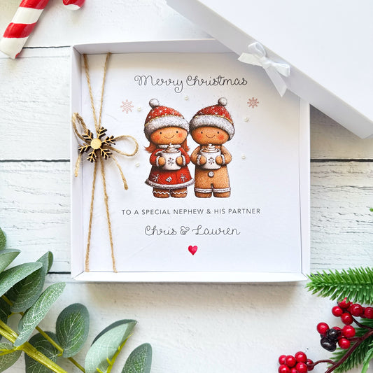 Personalised Christmas card for a couple featuring two cute gingerbread characters wearing red Santa hats and holding mugs of hot chocolate. The card reads Merry Christmas, with custom relationship wording and names Chris and Lauren. Hand-finished with rustic twine, a wooden snowflake and pearl embellishments. Shown with a Kraft envelope and optional white presentation box.