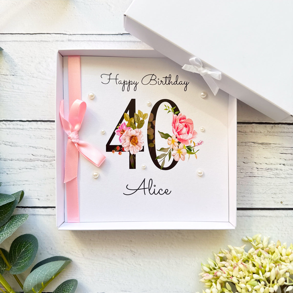 Personalised floral 40th birthday card for her featuring a large black number 40 decorated with pink roses and delicate flowers. The name Alice is printed beneath the number in elegant script. Hand-finished with a pink satin ribbon bow and pearl embellishments. Shown with a coordinating pink envelope and optional white presentation box.