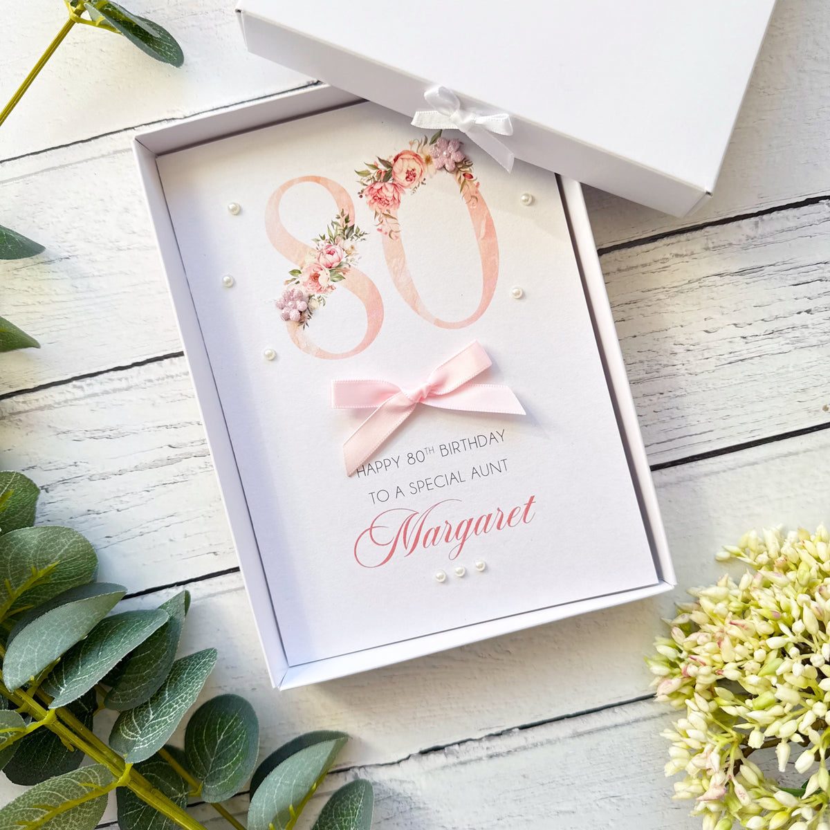 An open white gift box containing a personalised 80th birthday card for a special aunt named Margaret. The card features a large pink '80' decorated with pink and green floral arrangements, a light pink bow, and elegant script text.