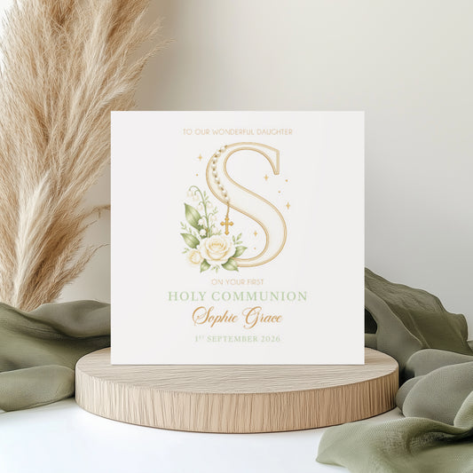 Elegant personalised First Holy Communion card featuring a floral monogram letter “S” decorated with rosary beads, a cross charm, white flowers, and soft greenery. The card includes personalised name and date details in delicate typography. Displayed on a neutral styled background with pampas grass and soft fabric accents.